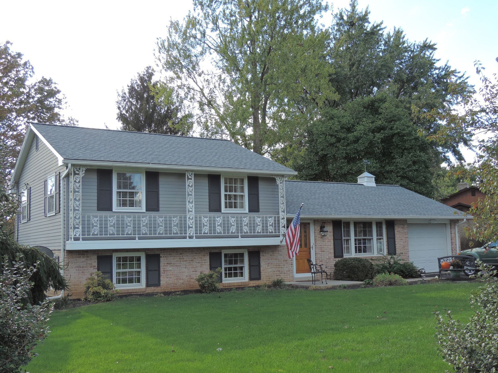 Split-level house with gray siding, brick, and a small porch; green lawn, trees, and an American flag.