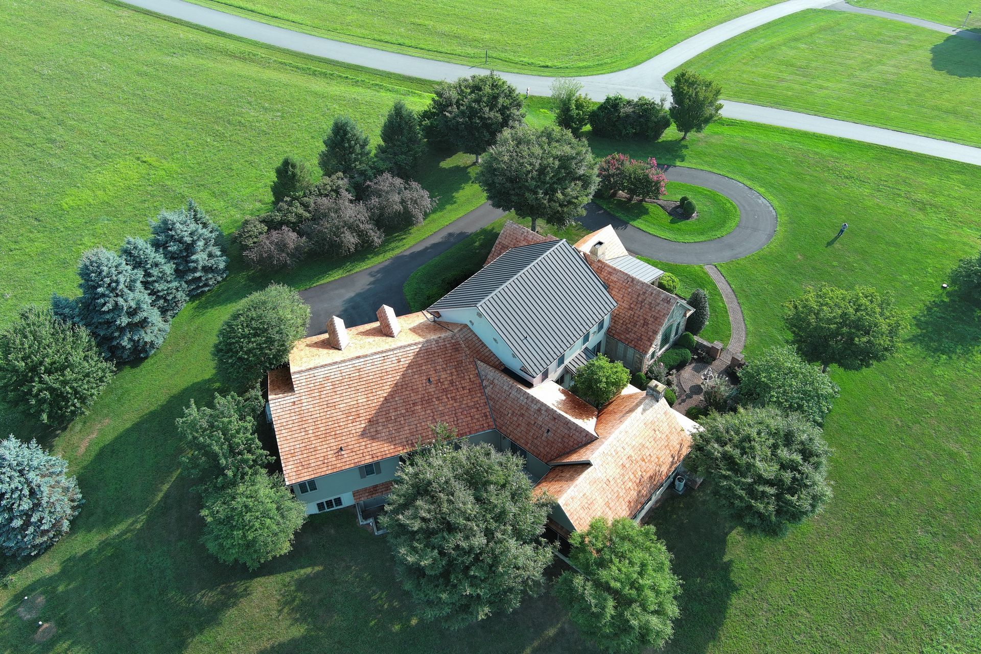 Aerial view of a large house with cedar roofs and a curved driveway, surrounded by trees and green fields.