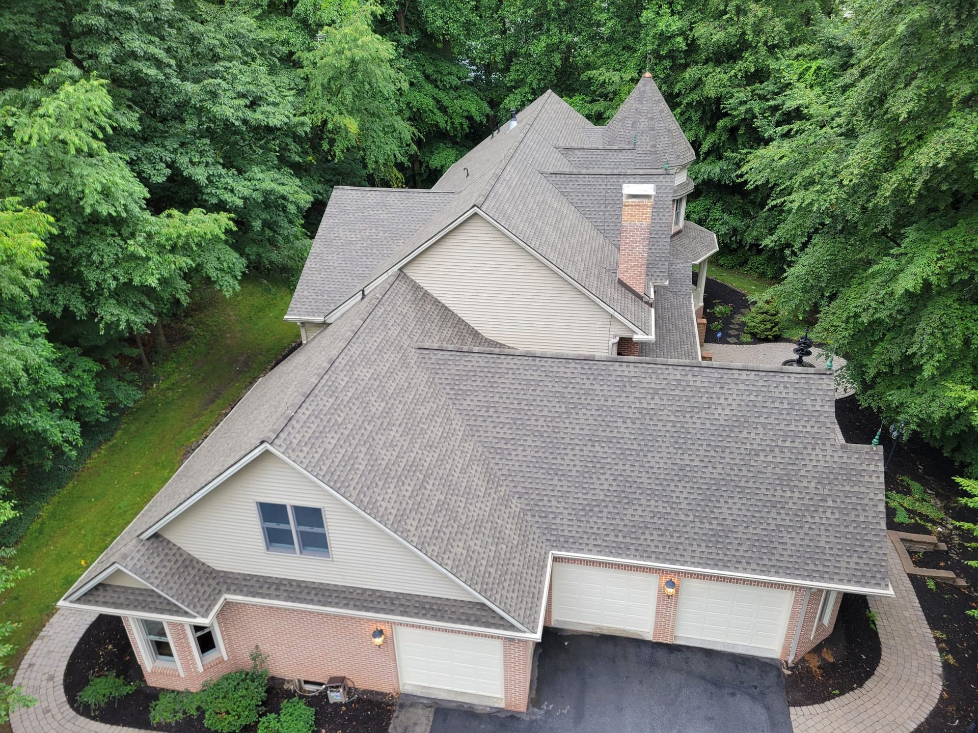 An aerial view of a multi-story house with a grey shingled roof, tan siding, brick accents, and a three-car garage.