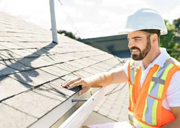 Roofer in hard hat and safety vest inspecting a residential roof, smiling in daylight.