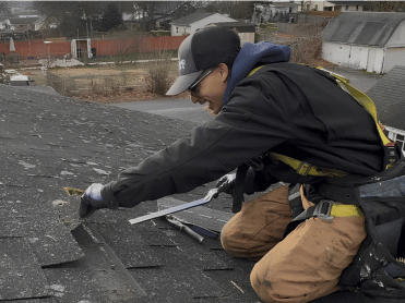 Roofer on a roof, kneeling, working with a tool. Wearing safety harness and gloves.