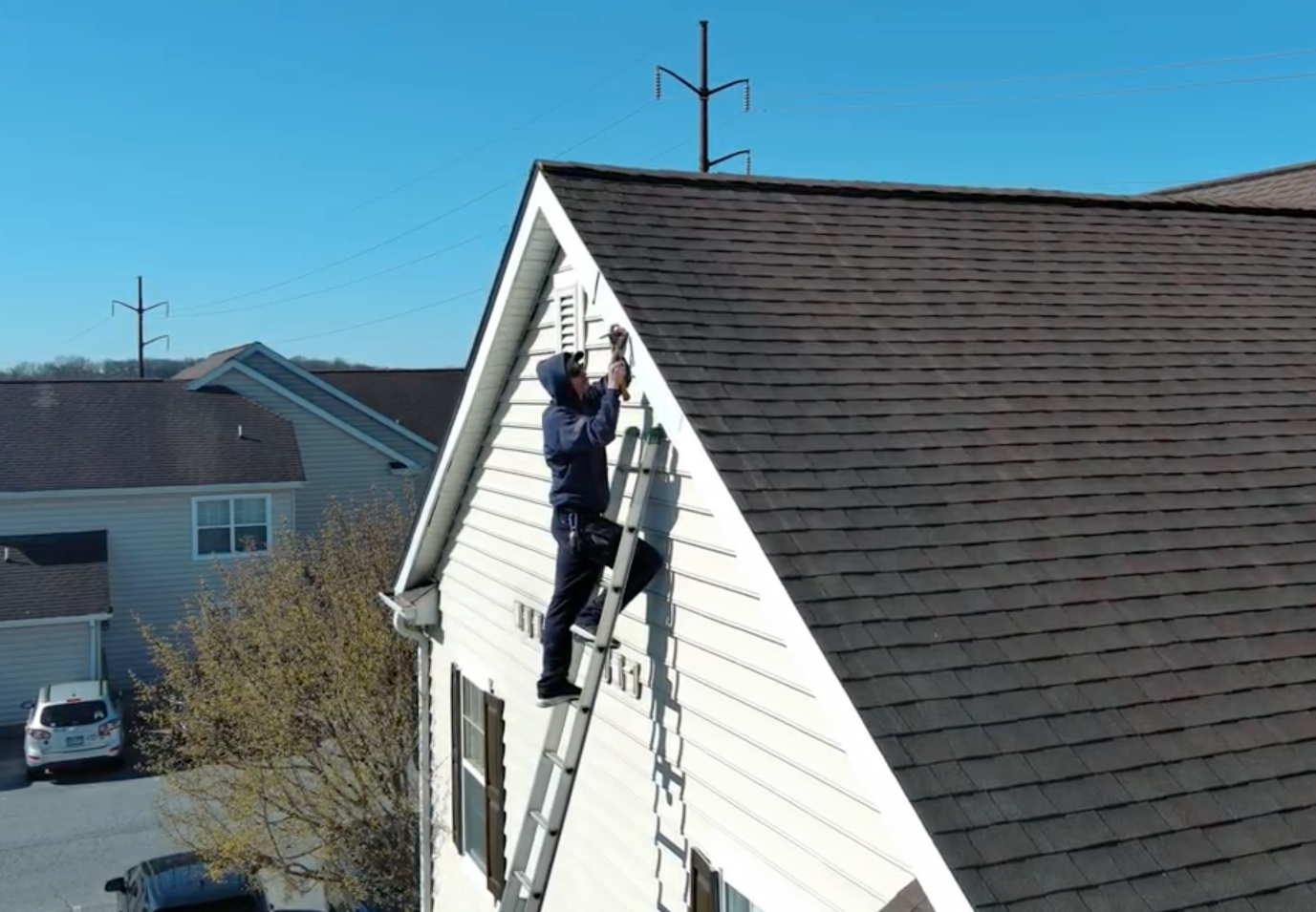 Person on ladder near a house roof, working on a utility box, bright sunny day.