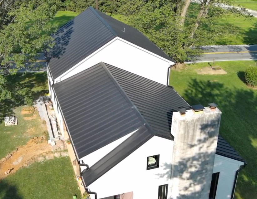 Aerial view of a white house with a black metal roof and brick chimney, surrounded by trees and lawn