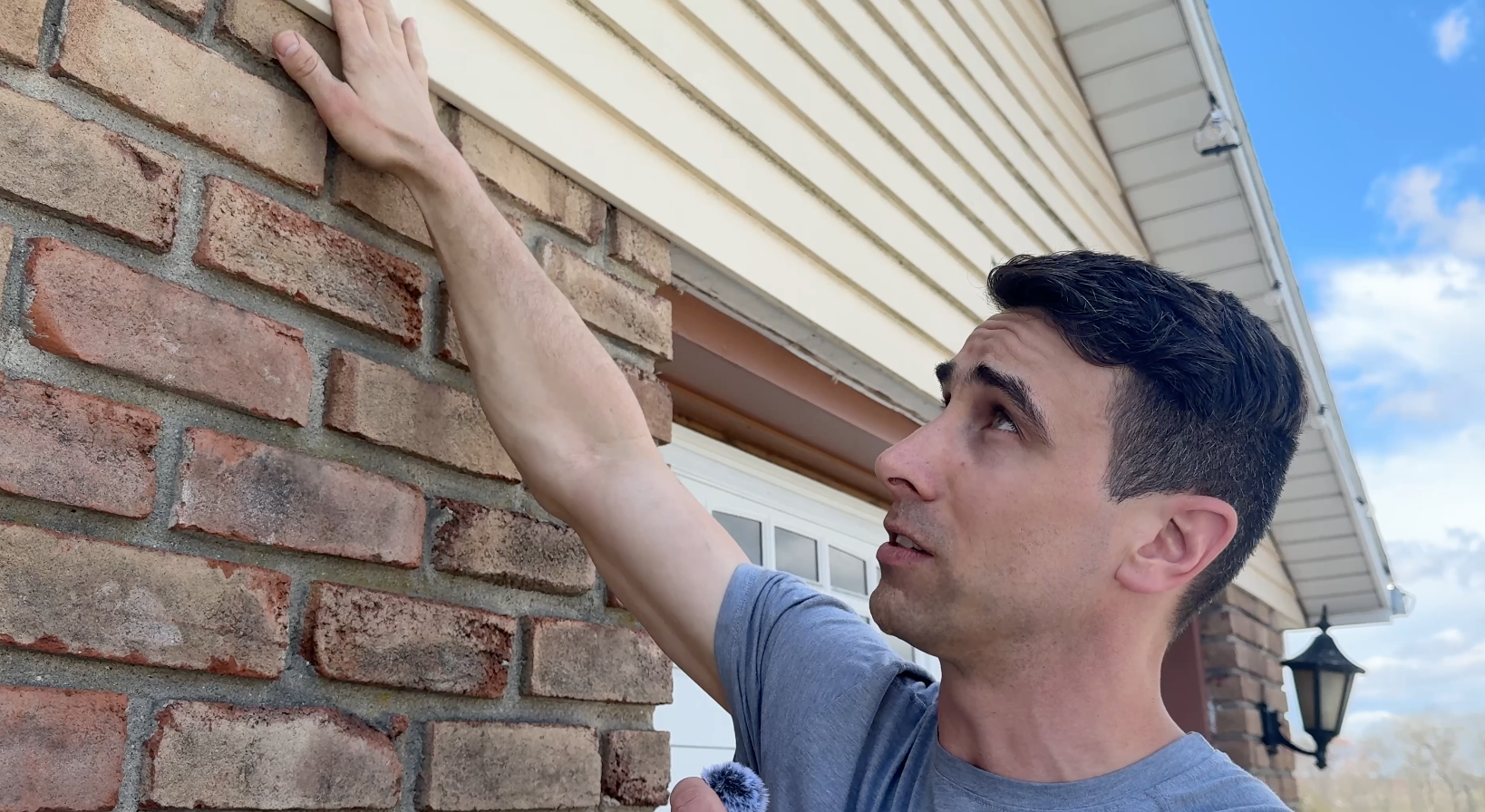 Man inspecting a brick wall and beige siding, reaching up near the roofline outdoors.