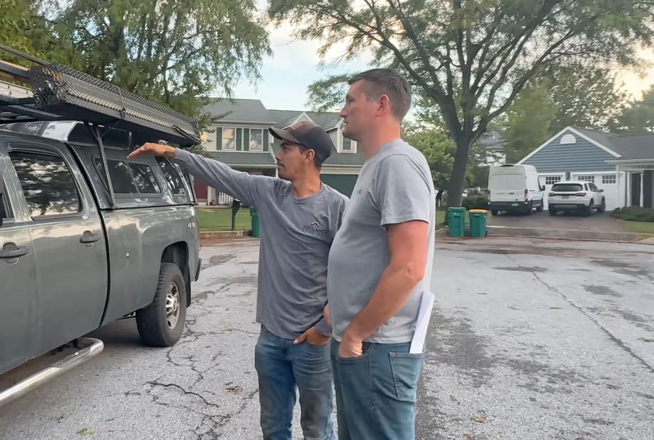 Two men stand by a dark pickup truck in a wet suburban parking lot, one pointing towards something off frame.
