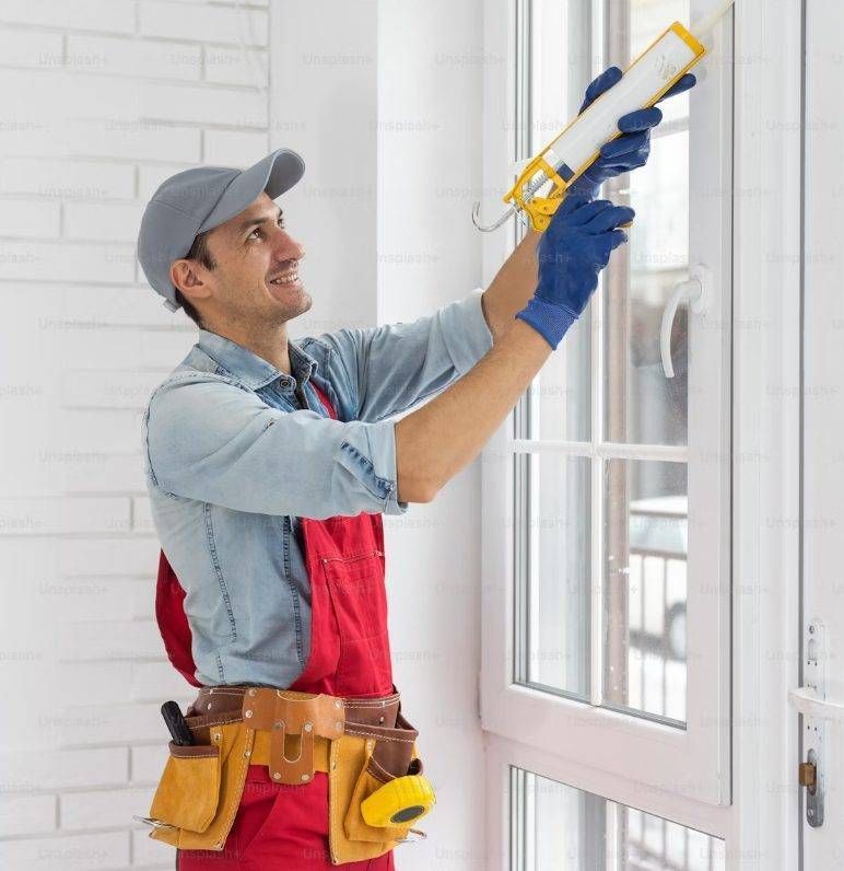 Man in work clothes uses a caulking gun on a window frame, inside a room.