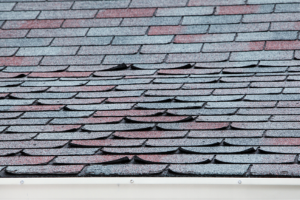 Close-up of a roof with buckled, discolored asphalt shingles in shades of blue, gray, and red.