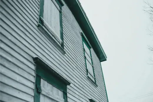 Side view of a weathered, light blue house with two boarded windows and a dark teal trim against a gray sky.