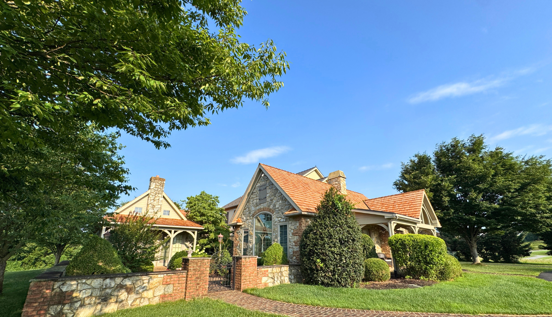 Stone cottage with red-tiled roofs and a small gazebo, surrounded by green trees and lawn under a blue sky