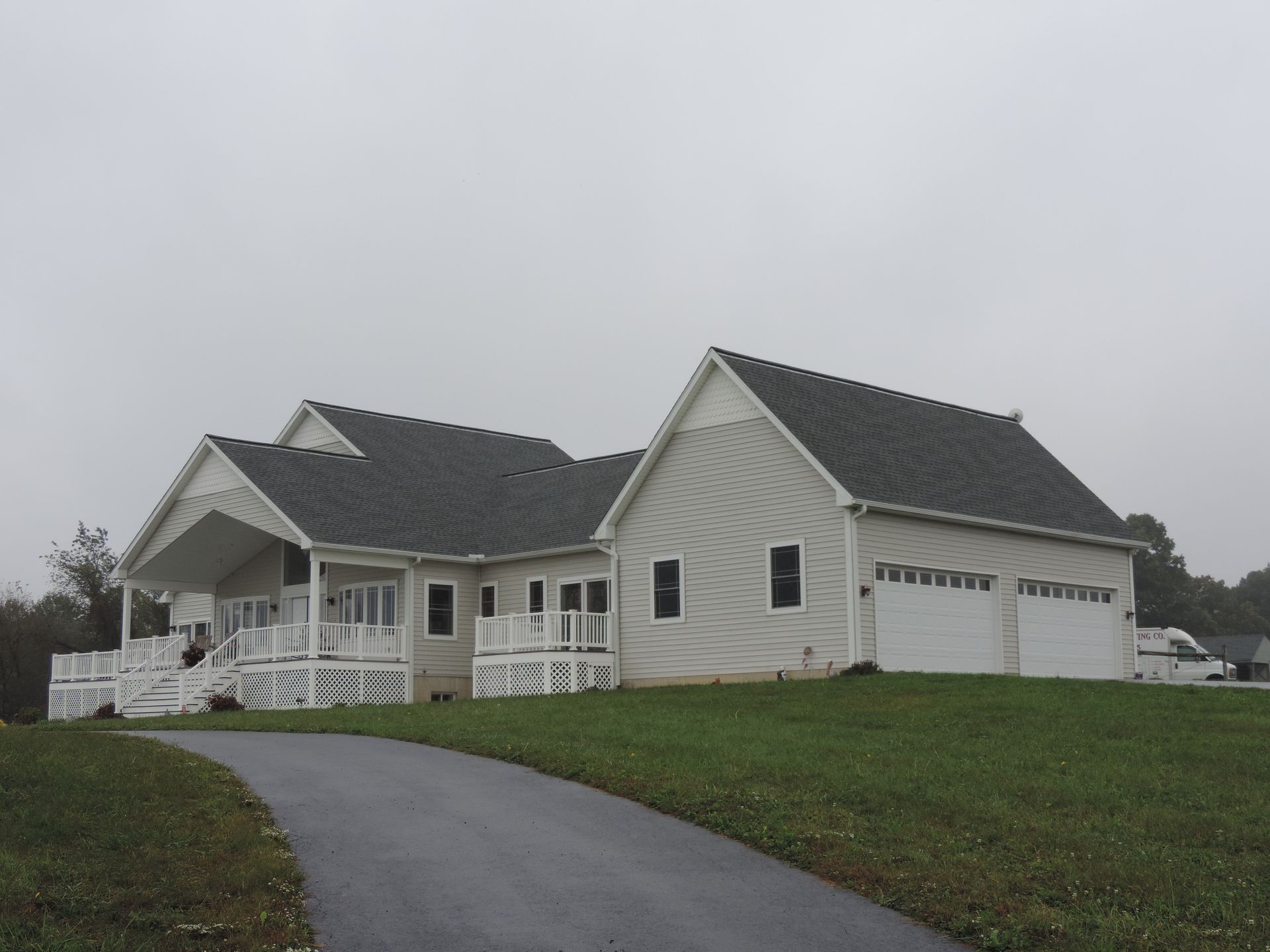 White house with attached garage and a curved driveway on a grassy hill under an overcast sky
