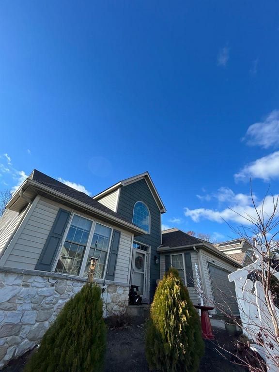 Stone house with gray trim and dormer windows under a bright blue sky, framed by shrubs in front.