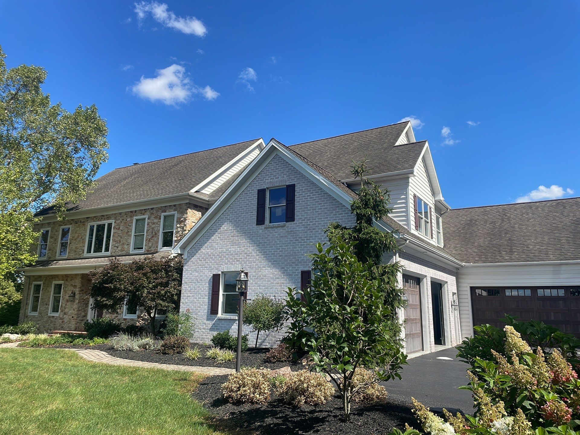 Large gray suburban house with a stone facade, garage, and manicured lawn under a blue sky