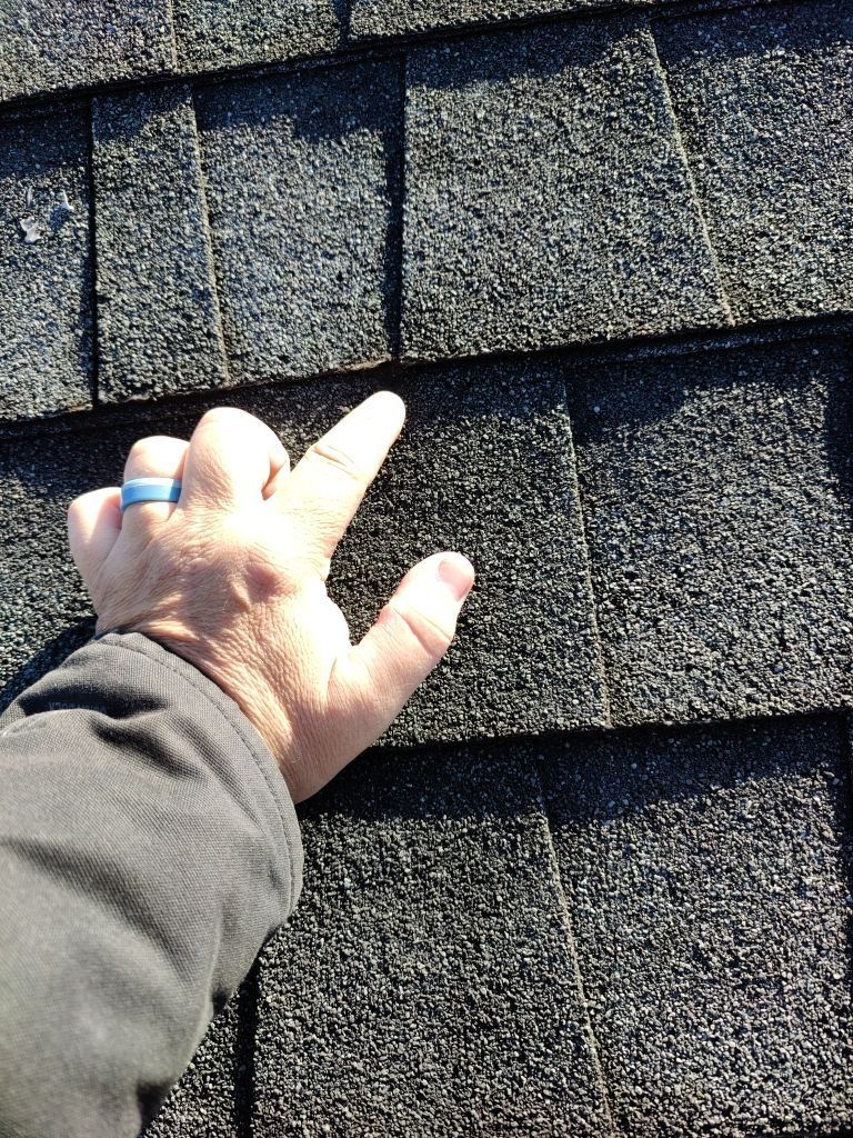Hand checking gray asphalt roof shingles for damage, with one shingle lifted.