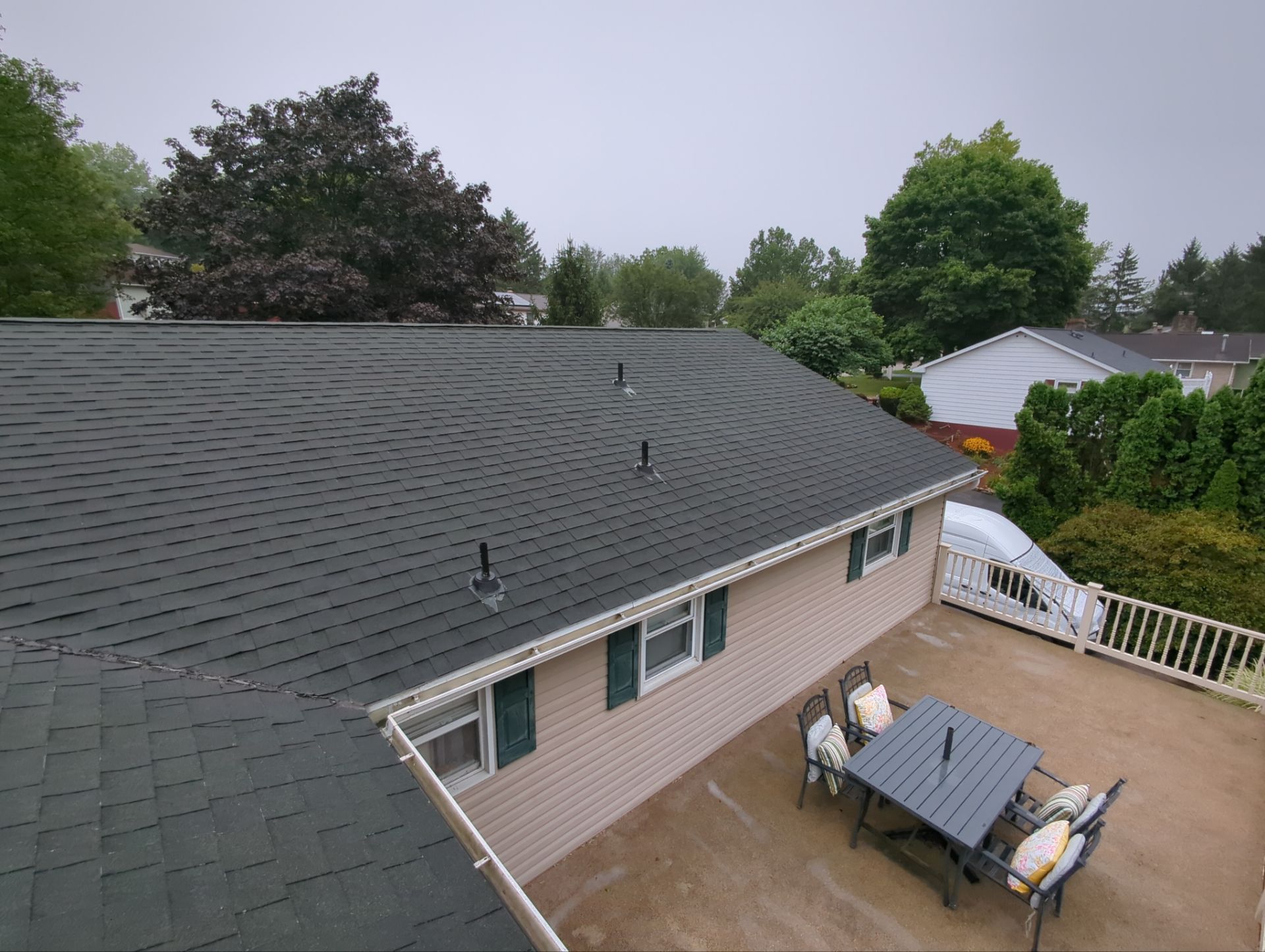 Backyard patio beside a gray shingled roof, with outdoor table, chairs, and surrounding trees.
