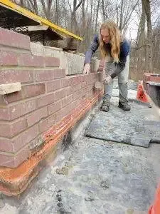 Person building a brick wall outdoors, using a trowel to apply mortar.