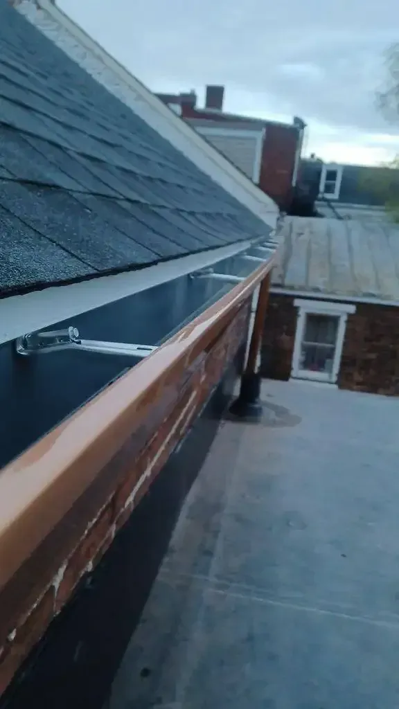 Copper gutters along a dark shingle roof edge, with a glimpse of neighboring buildings.
