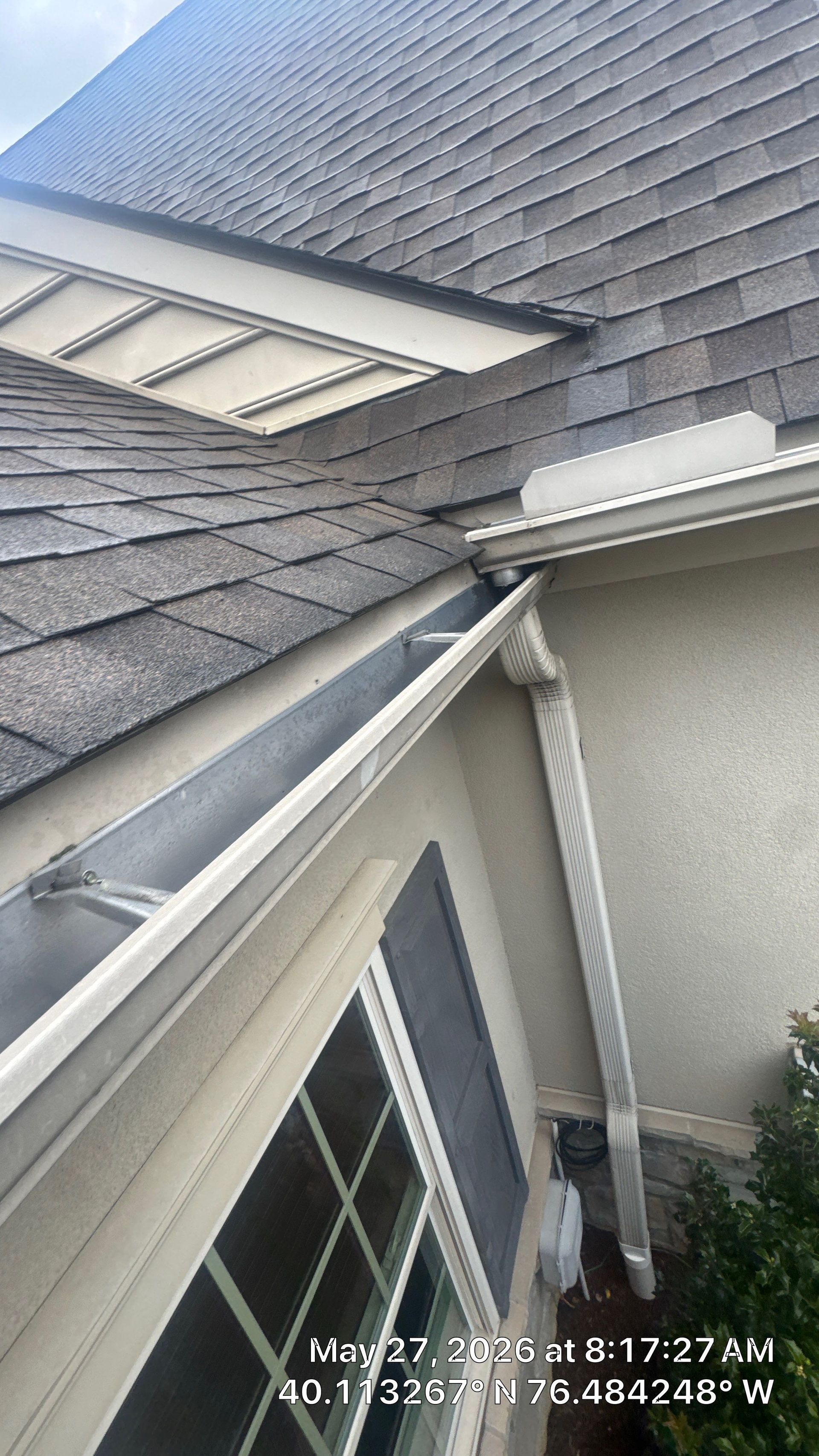 Rain gutter and roof edge beside a sloped shingle roof and window on a house exterior.
