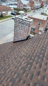 Chimney on a brown shingle roof, view overlooking a street with buildings and parked cars.