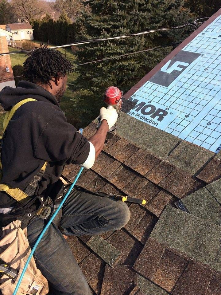 Roofer on a roof, using a nail gun to install shingles. Blue underlayment visible, sunny day.