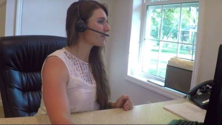 Woman wearing a headset, seated at a desk, looking at a computer screen next to a window.