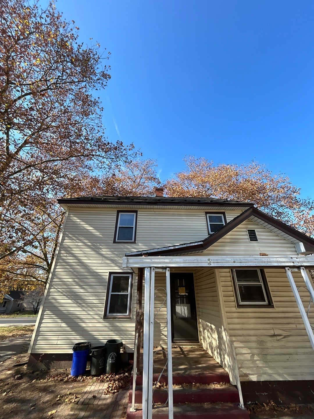 Small beige house with red steps, front porch, and autumn trees under a clear blue sky