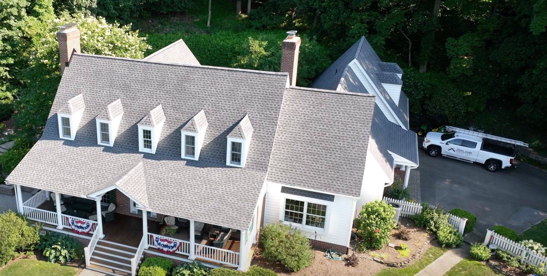 Aerial view of a gray-roofed house with a porch, dormer windows, and a service truck in the driveway.