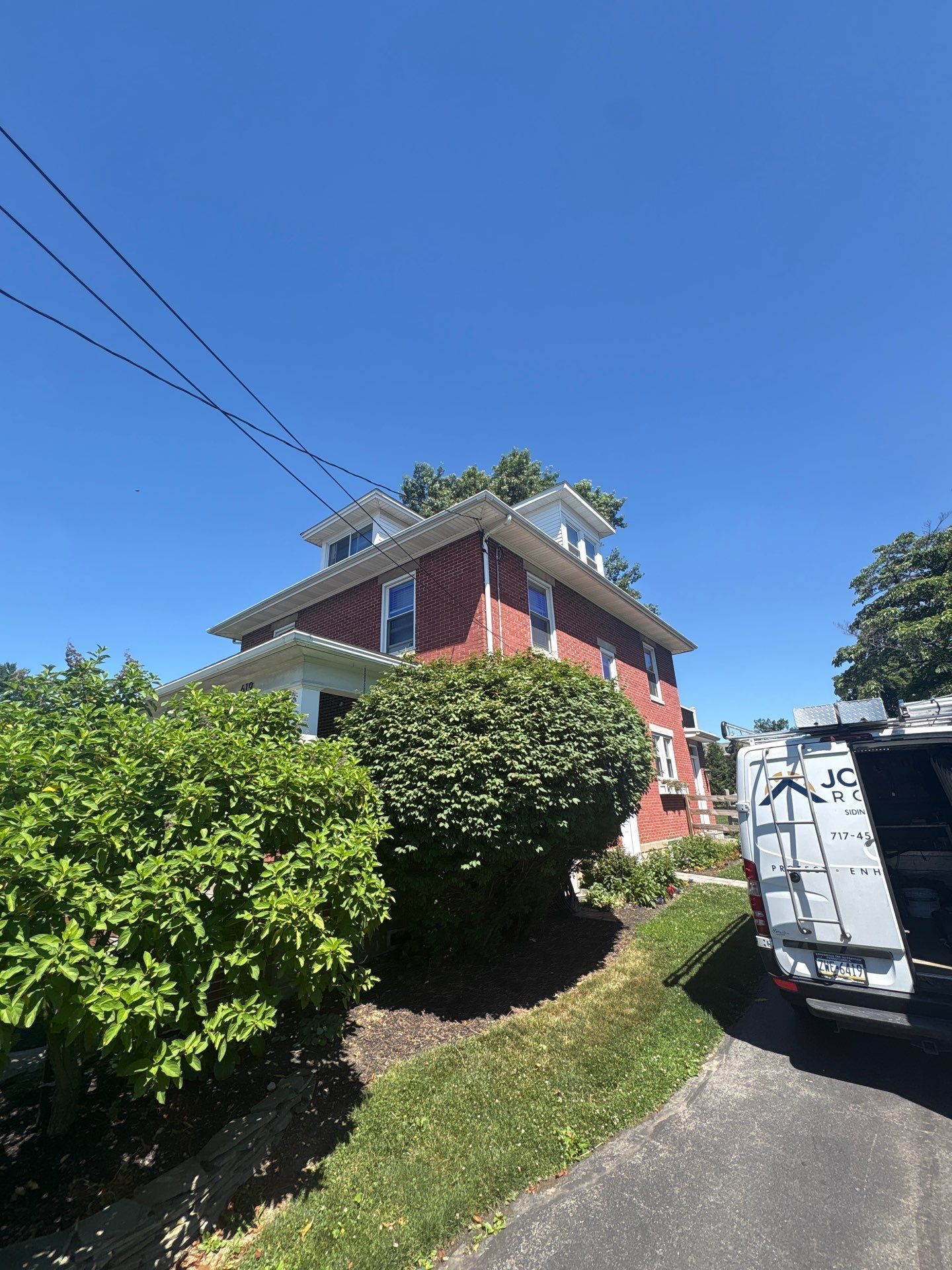Red house on a sloped road with green shrubs, blue sky, and a white van parked at right.