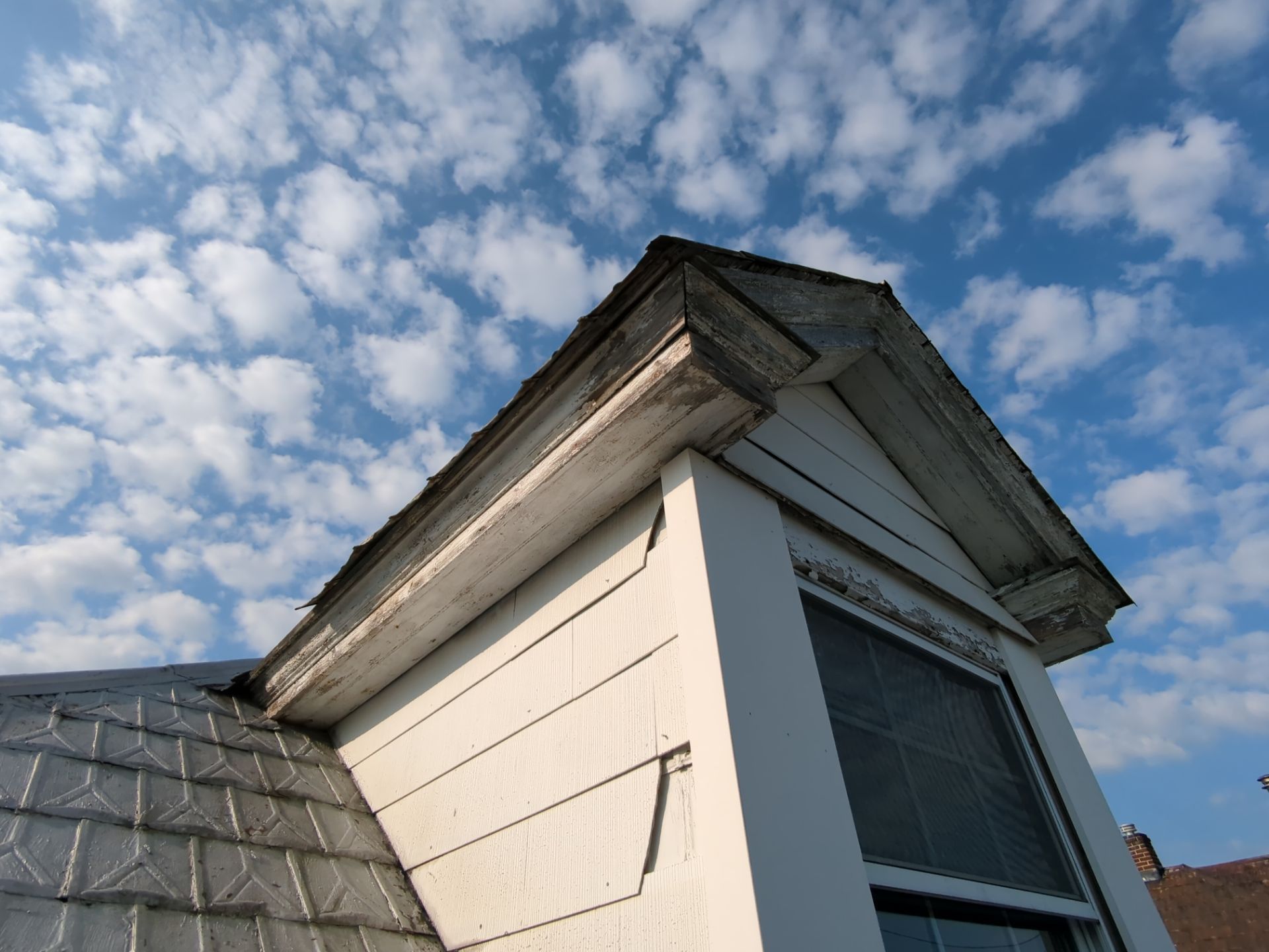 Dormer on a residential roof with peeling paint and deteriorated wooden fascia and trim during an exterior inspection.