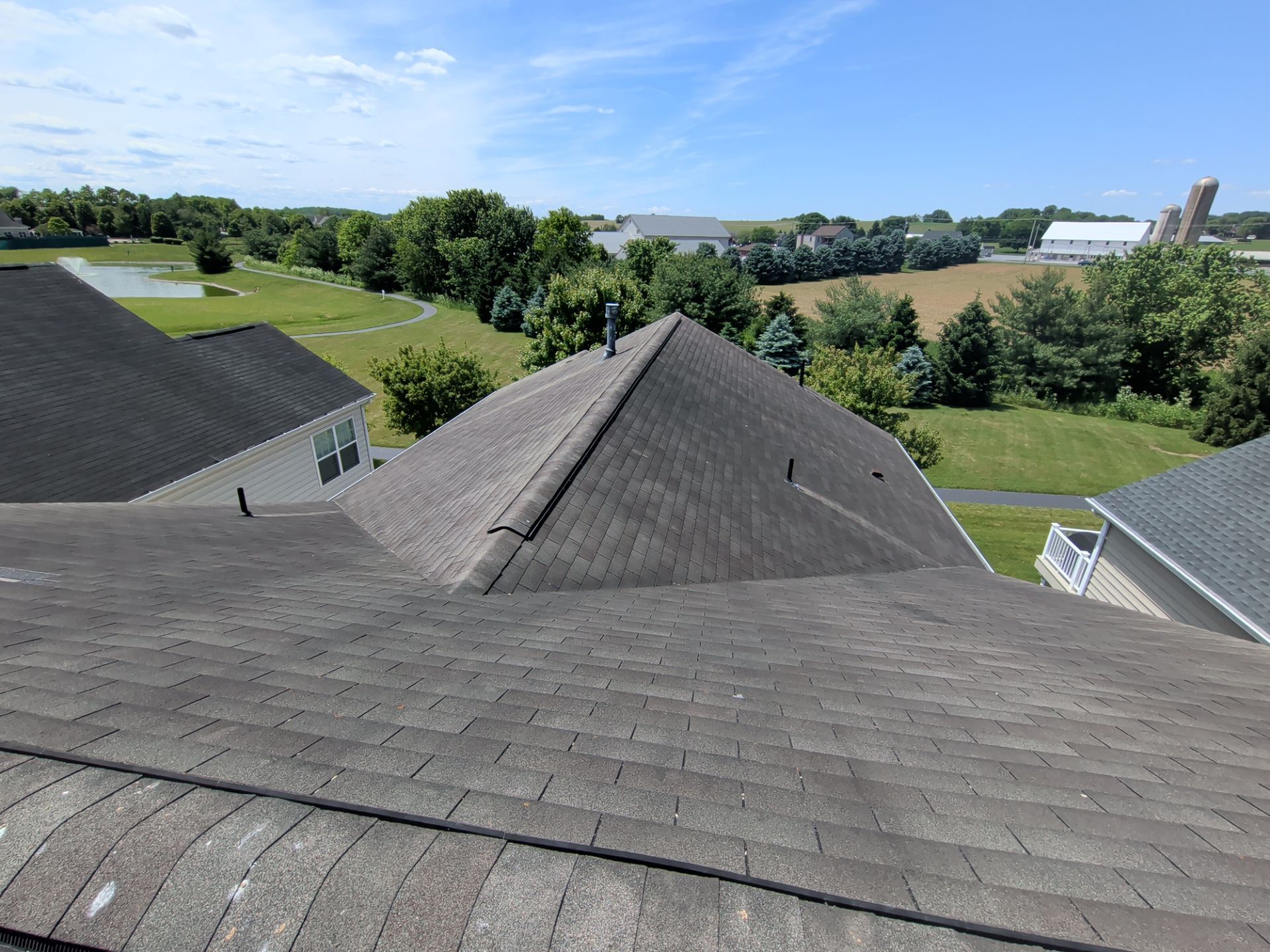 View from a rooftop over gray shingles to green fields and trees on a sunny day.