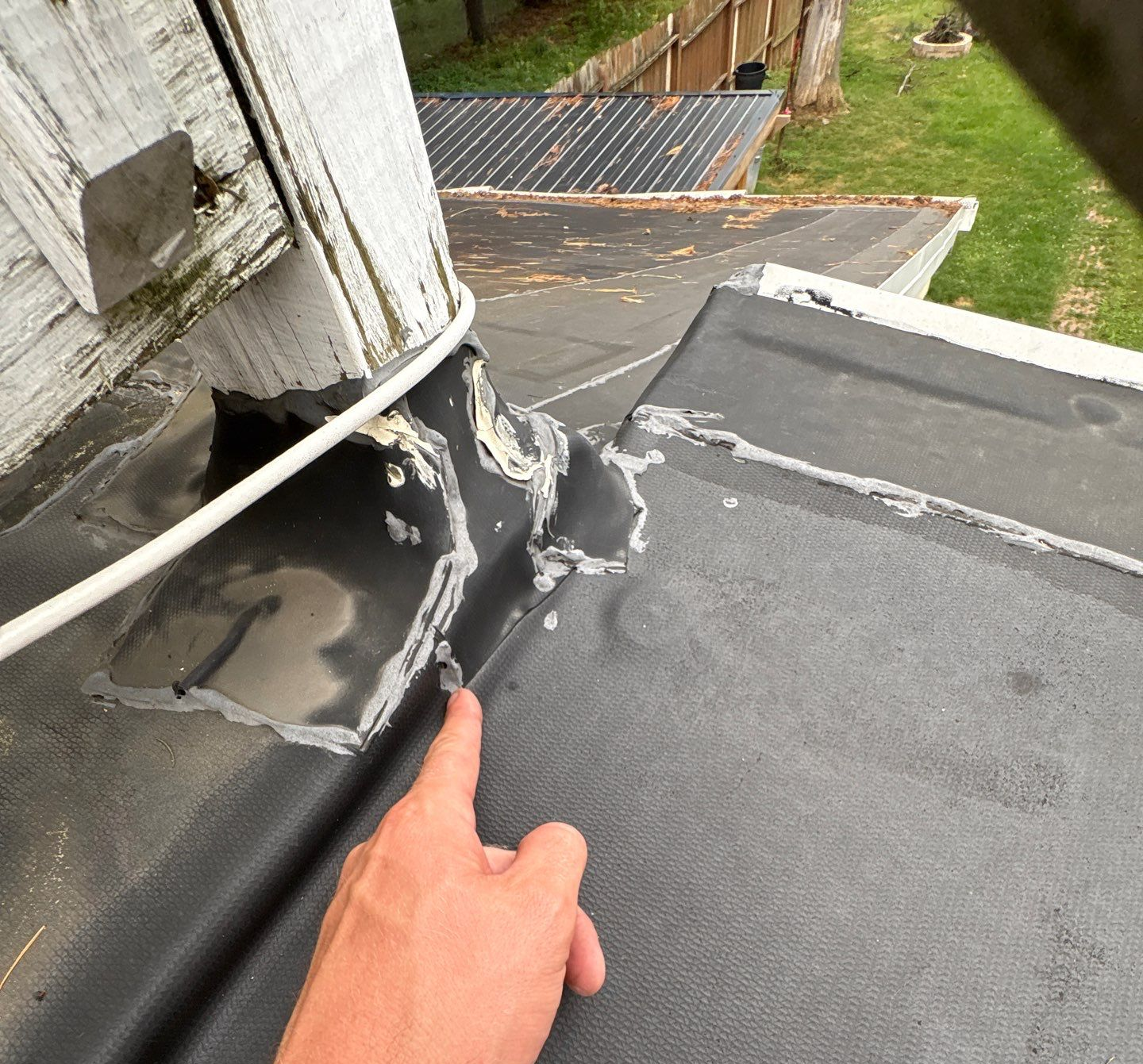 Hand pointing to damaged roof flashing and cracked sealant beside a chimney on a flat roof