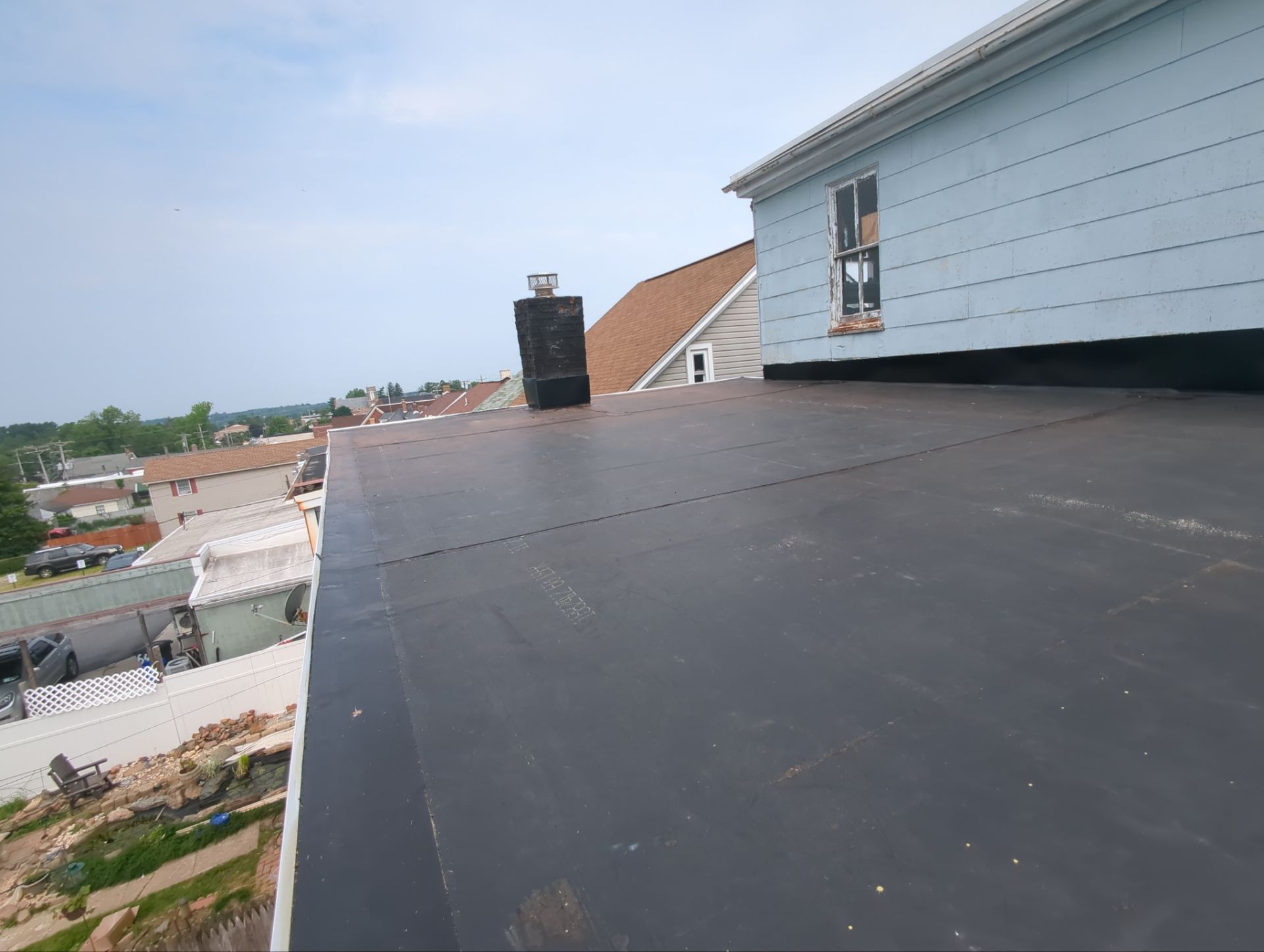 Flat black rooftop beside a light blue house with a chimney, overlooking a neighborhood below