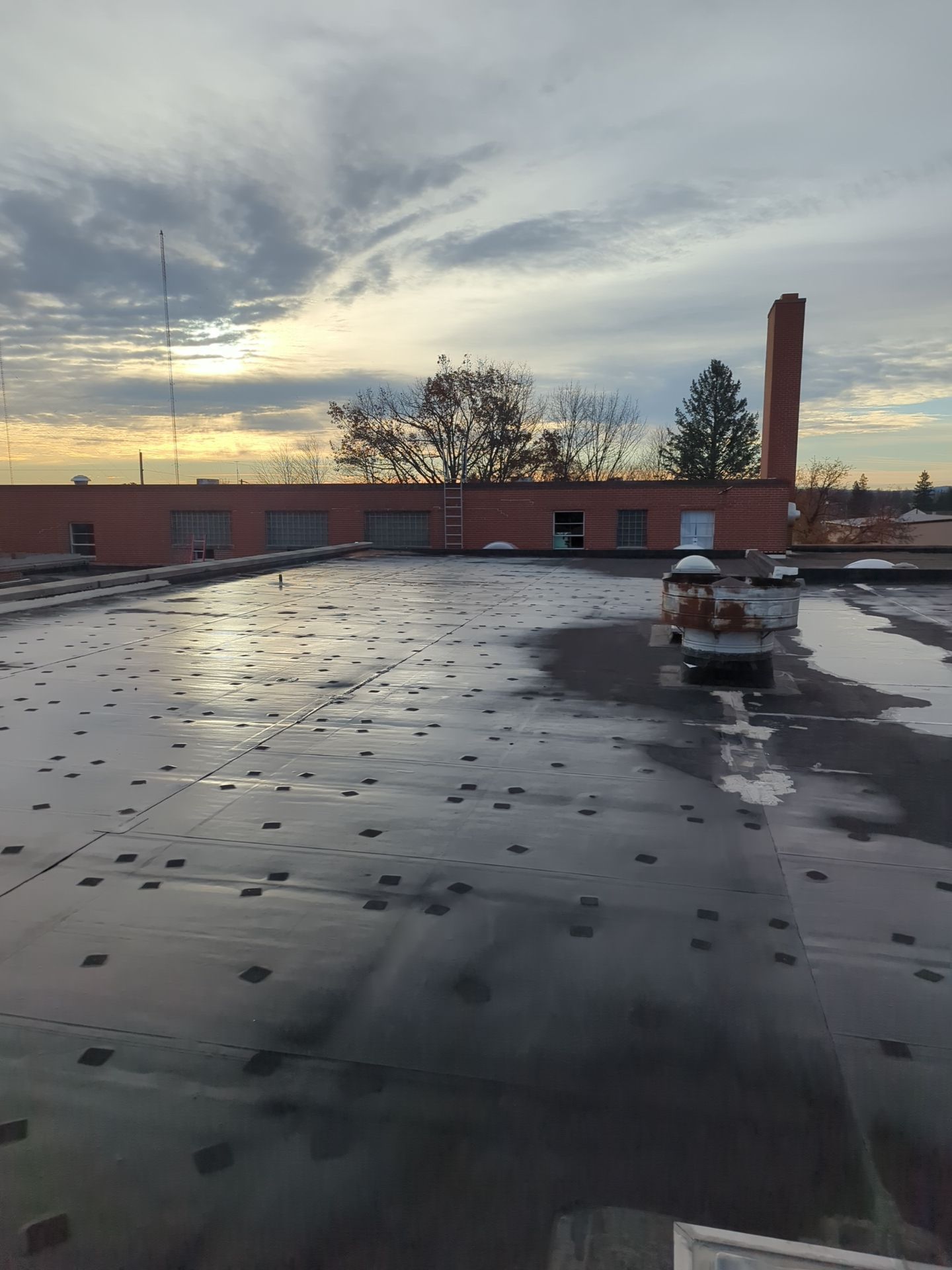 Rooftop at sunset with wet black roofing, a brick parapet, and a tall smokestack in the distance