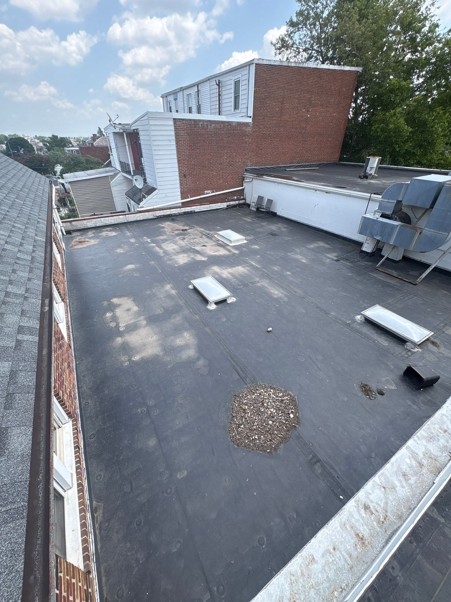 Rooftop with flat black roofing, white vents, and a damaged red-brick wall beside trees and nearby houses