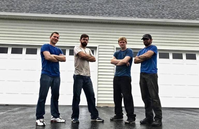 Four men standing with arms crossed in front of a white garage door. Blue and tan shirts, dark pants.