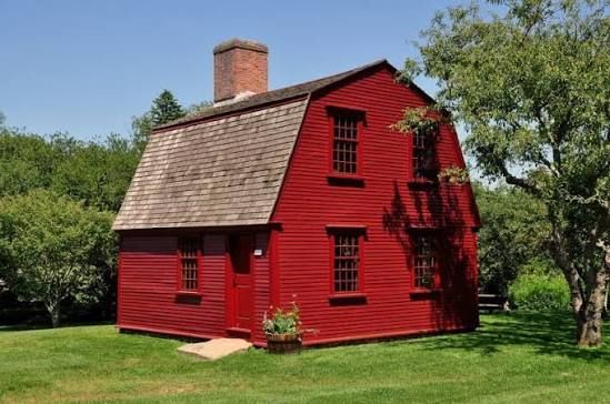 Red wooden house with chimney on a grassy lawn surrounded by trees.