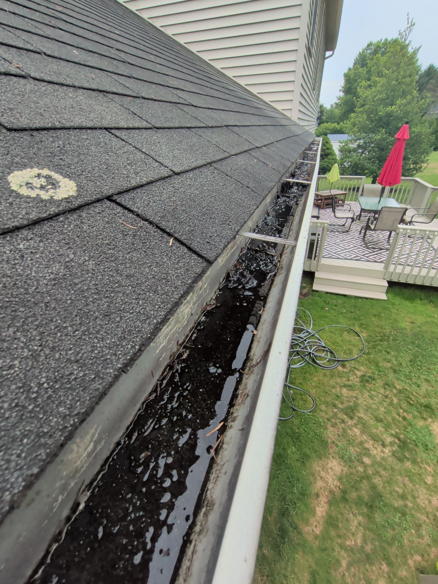 Close-up of a roof gutter overflowing with debris beside a house and backyard patio.