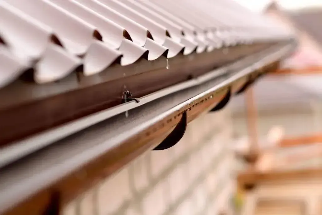 Close-up of a corrugated roof edge with water dripping off, set against a blurred brick wall.