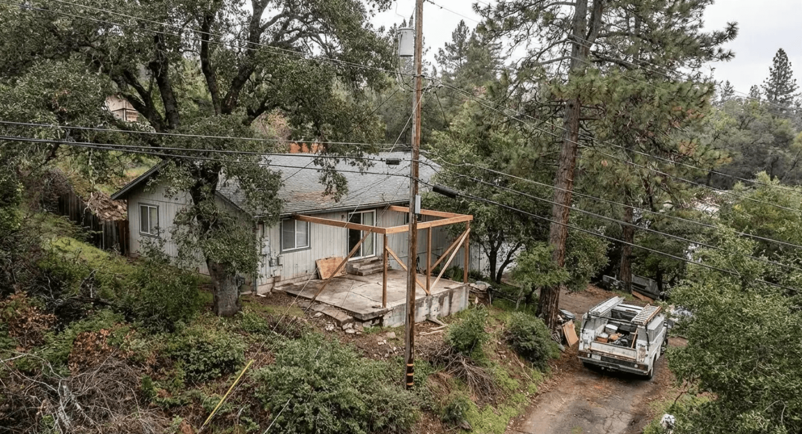 Patio roof framing under construction on a heavily sloped lot surrounded by trees, where site complexity drives costs well beyond standard patio roof ideas