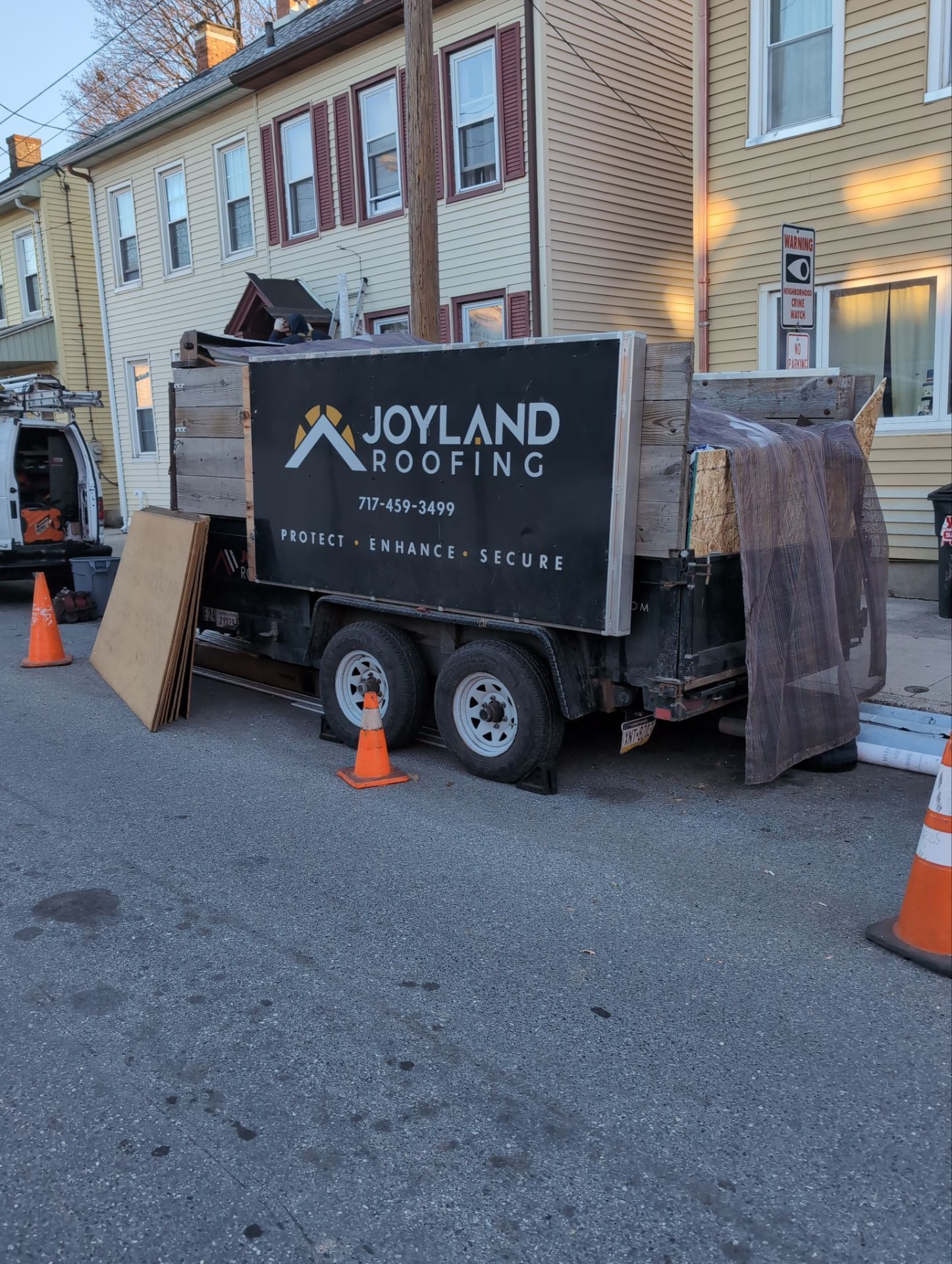 Joyland Roofing truck parked on a residential street beside rowhouses, with orange cones and a ladder nearby