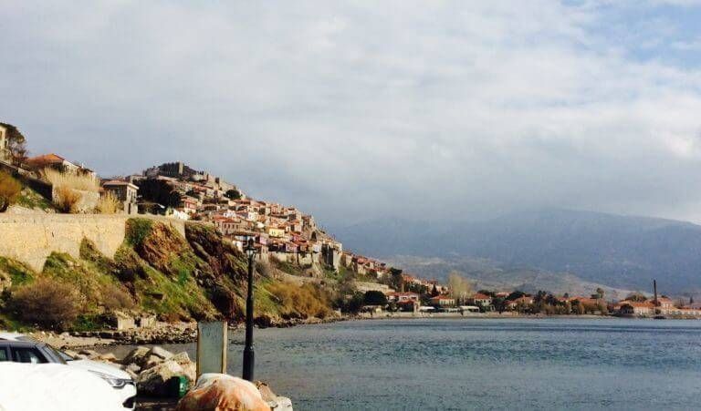 A town with red-tiled roofs sits on a green hillside by a calm lake, under a cloudy sky.