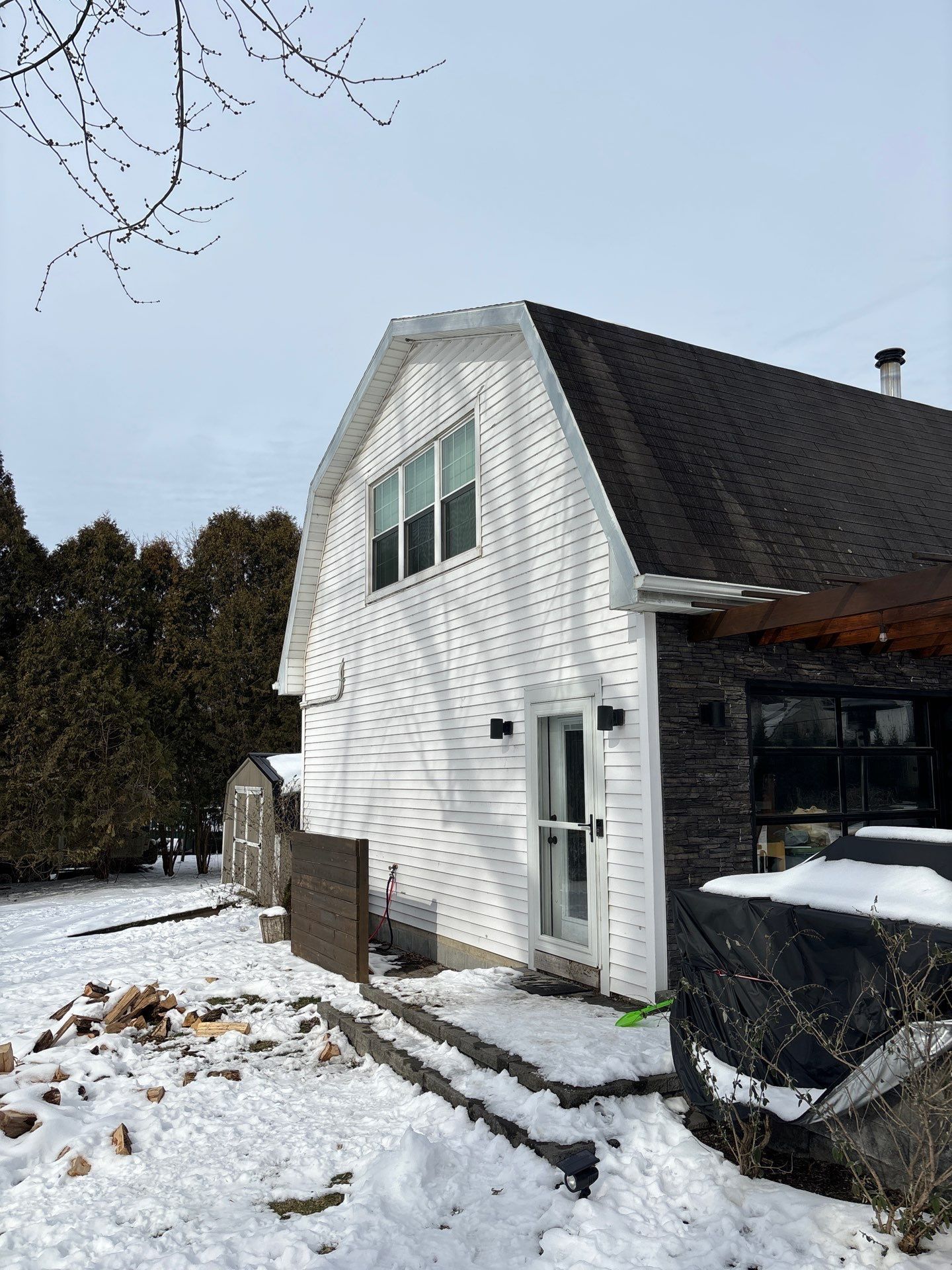 Snow-covered white house with dark roof and bare trees beside a driveway