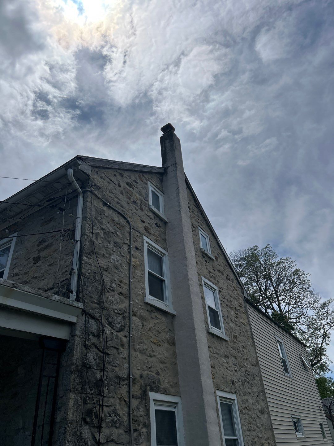 Stone house corner under a cloudy sky, with a tall chimney and white-trimmed windows.