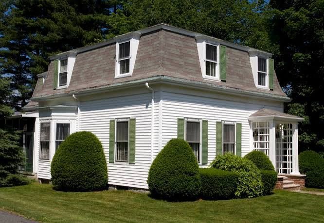 White colonial house with dormer windows, green shutters, and manicured shrubs on a sunny lawn