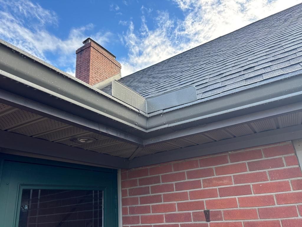 Low-angle view of a red brick house corner showing a roof eave, dark gutters, and a brick chimney against a blue sky.