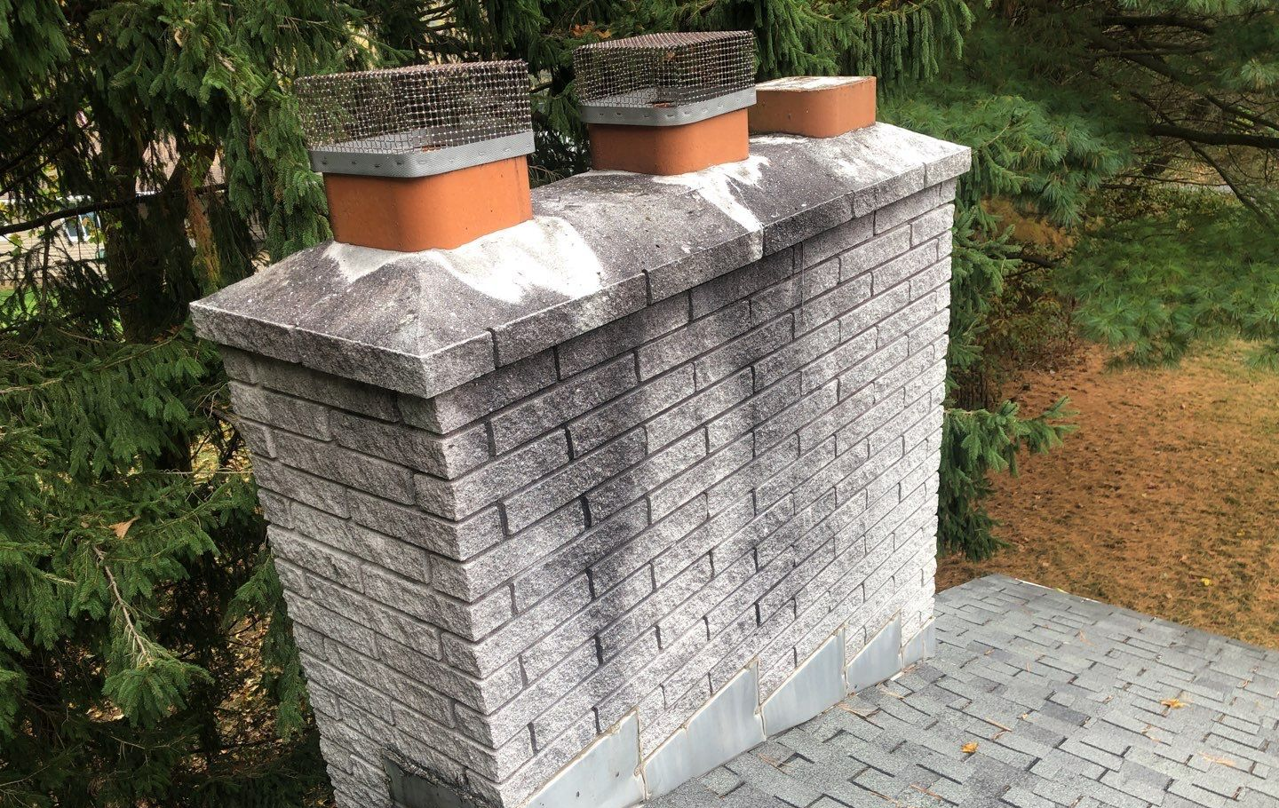 Brick chimney with three orange-capped flues on a shingled roof, surrounded by trees and greenery