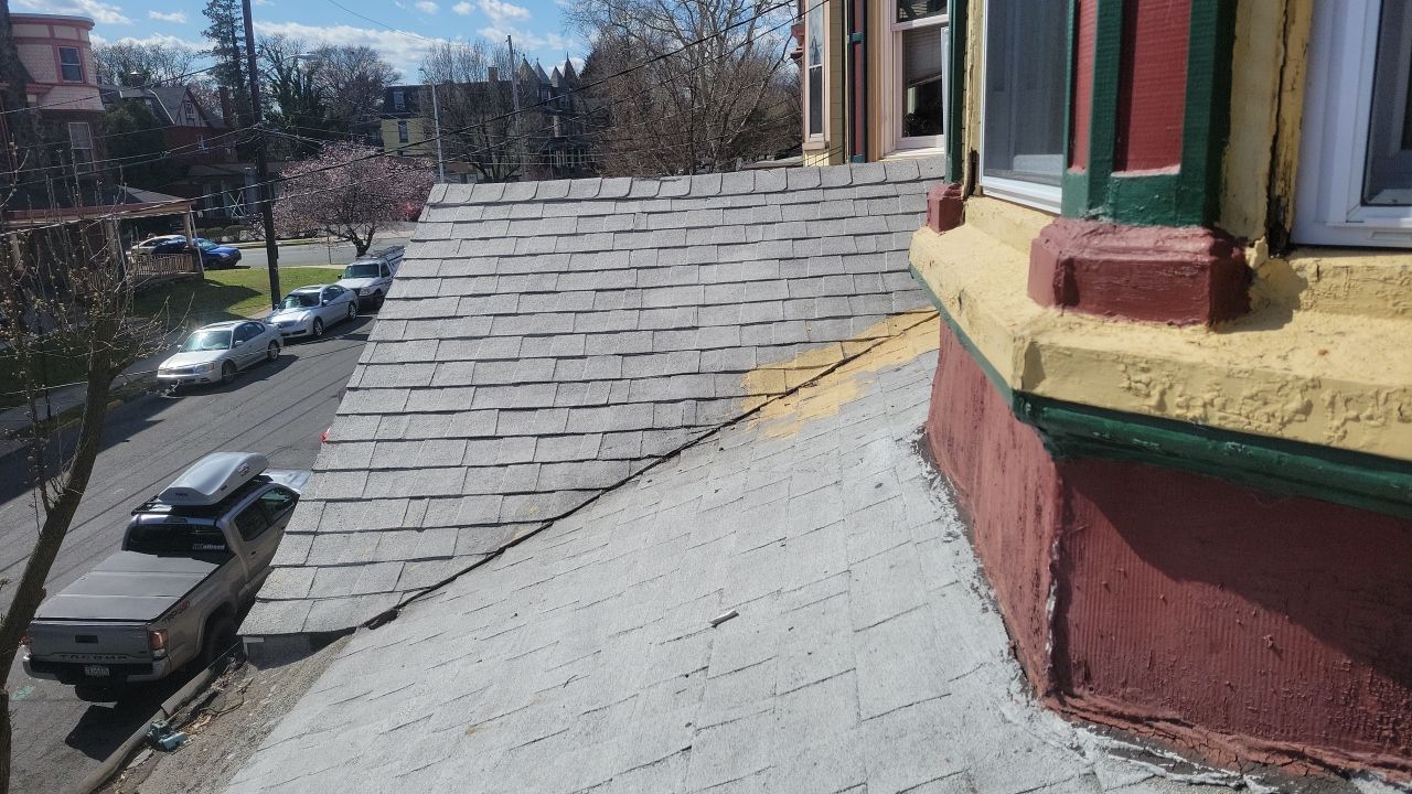 Rooftop with flat black roofing, white vents, and a damaged red-brick wall beside trees and nearby houses