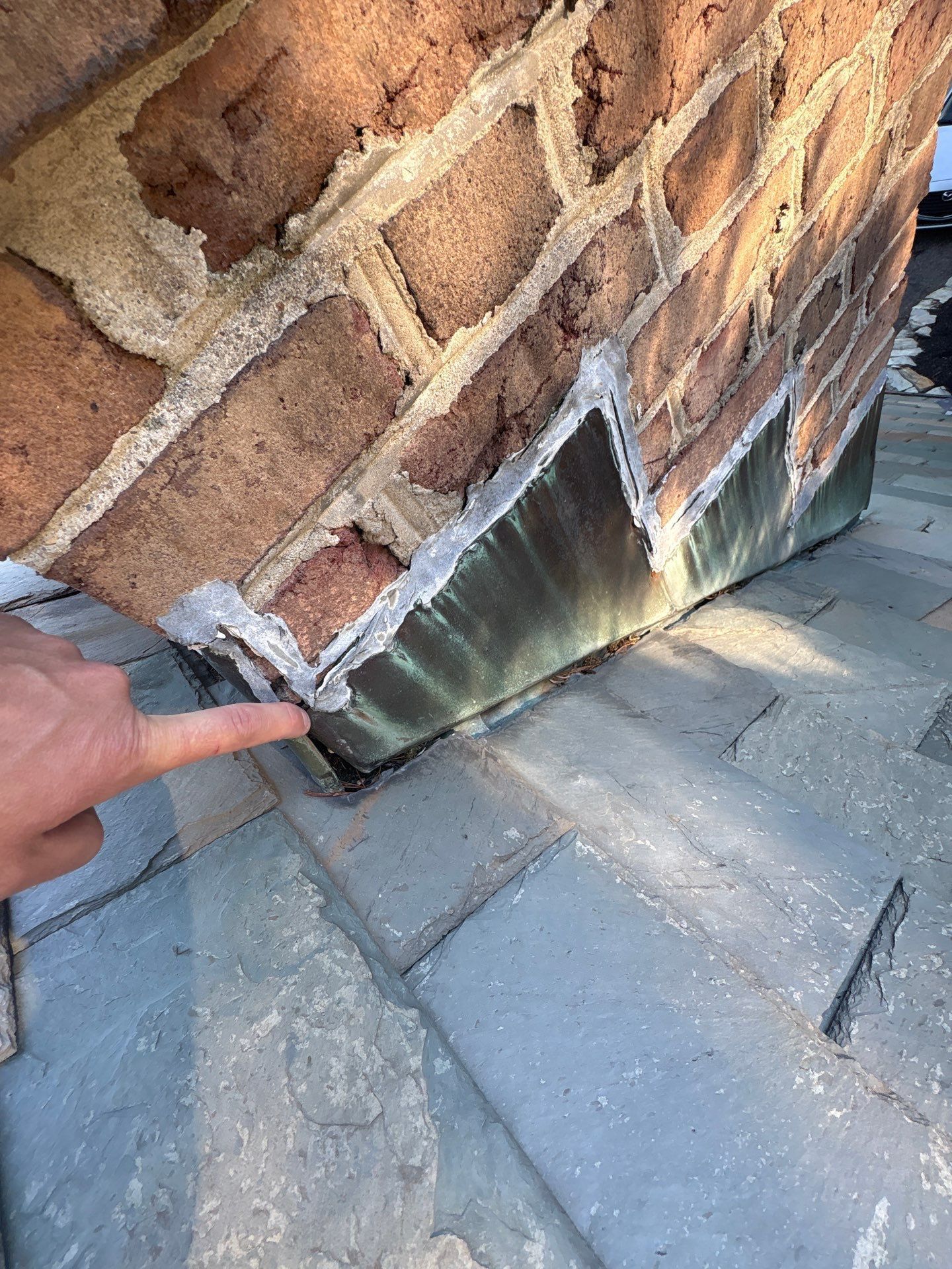 Finger pointing at a damaged brick wall and flashing metal at the base near a pavement.