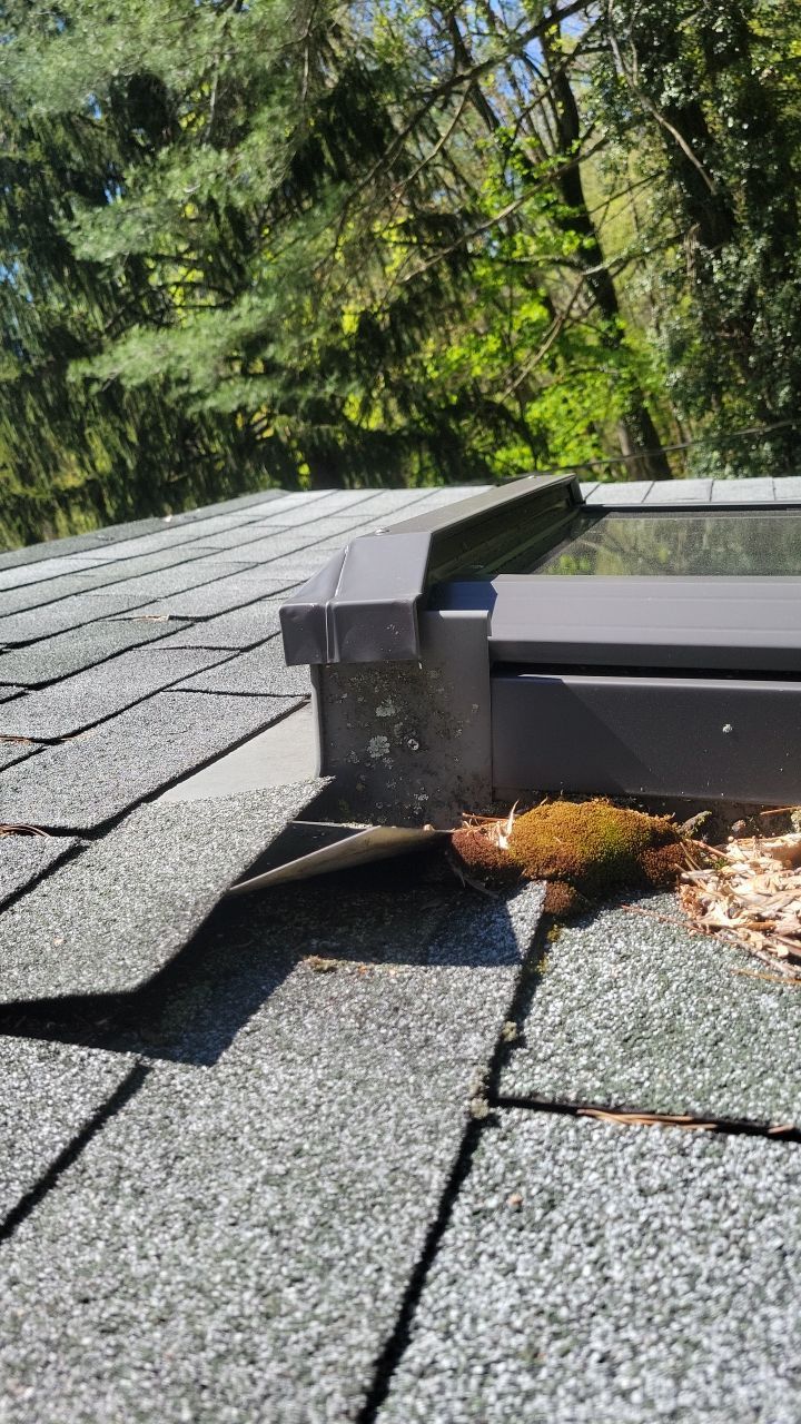 Damaged roof shingles around a black chimney flashing, with debris and trees in the background