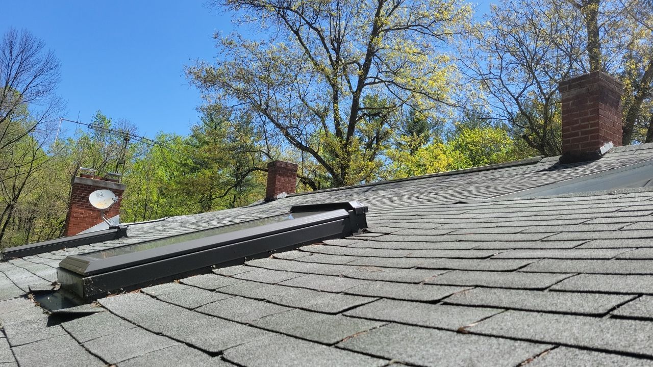 Roof shingles with a dark skylight and two brick chimneys against a blue sky and trees