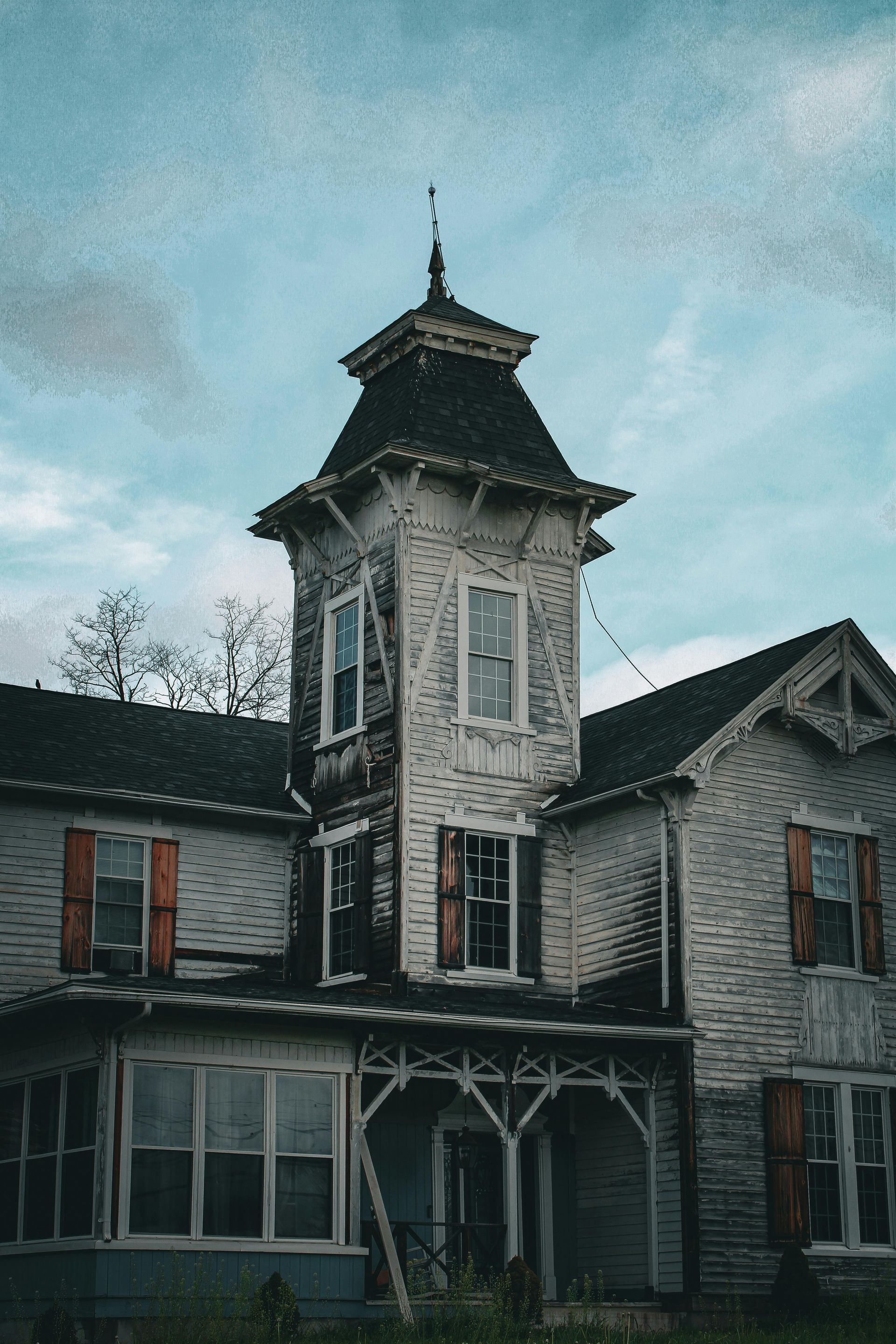 Old Victorian house with a central tower and weathered white siding under a cloudy blue sky